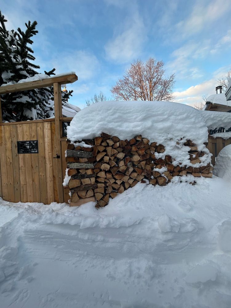 A five foot high wood pile topped with a couple of feet of snow and buried at its base with the same. 