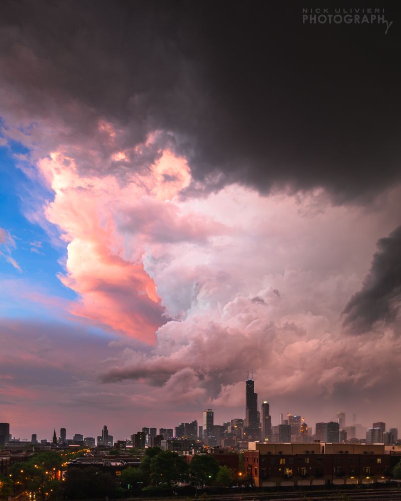 A dramatic thunderstorm looms over the Chicago skyline at sunset. The towering cumulonimbus is lit pink and orange by the setting sun on the left side, with a patch of blue sky peeking through. Meanwhile, darker, ominous clouds dominate the right side of the frame. The city below is partially shrouded in rain, with streetlights glowing in the foreground as twilight sets in.
