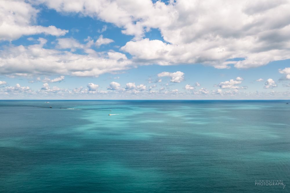 A drone view of an expansive Lake Michigan. White cumulus clouds fill the blue sky. The lake below is various shades of blue and turquoise the darker patches of lake are cloud shadows while the bright spots are where the sun is breaking through. A boat in the distance looks tiny as it's illuminated by the sun in Chicago