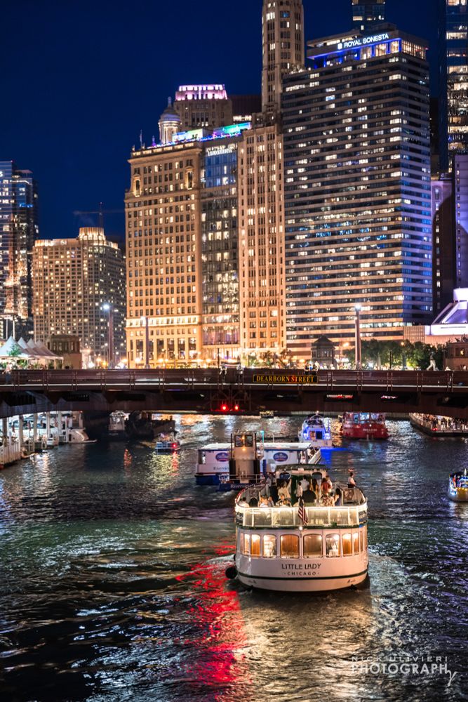A nighttime image of the chicago river at night. A boat passes in the foreground while the skyline of buildings along upper wacker are illuminated in and out from the city lights