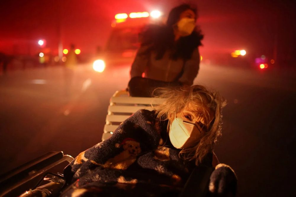 A woman in a mask sits in an improvised wheelchair and is evacuated by another woman behind her. In the background, the sky is colored orange by fires and smoke. Photo: Ethan Swope