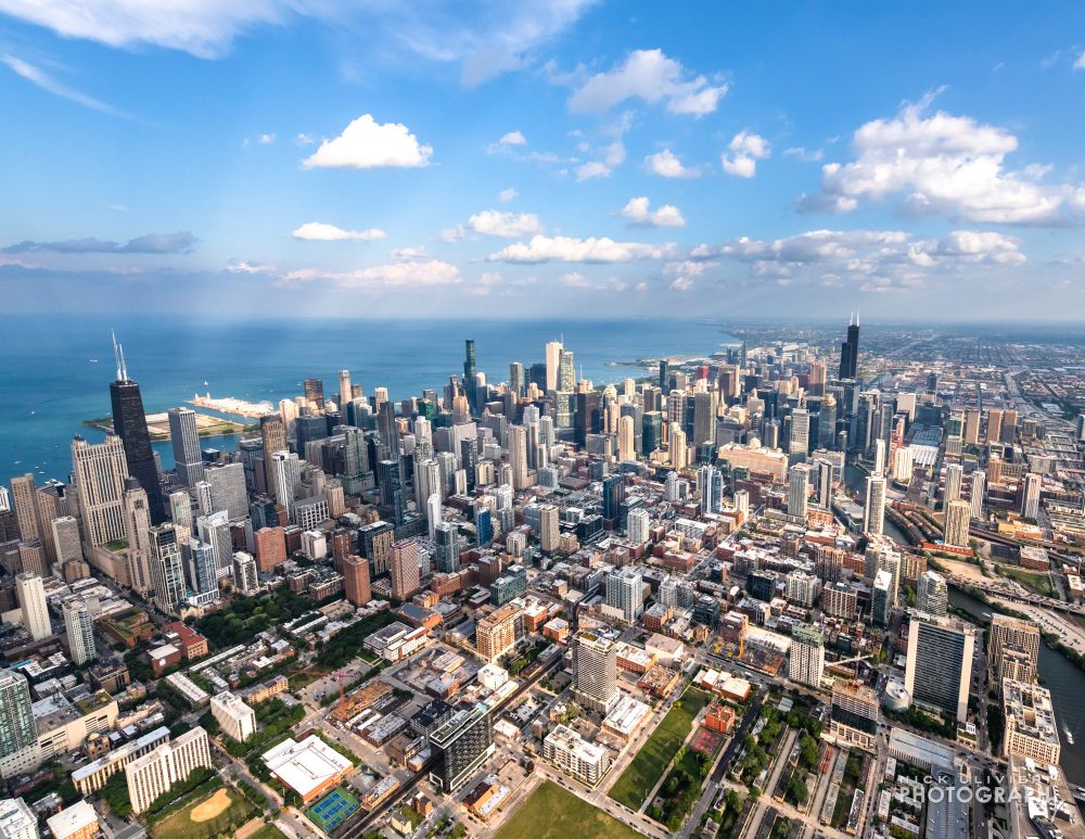 A view of Chicago from a helicopter on a sunny, summer day. The skyline is bathed in light with Lake Michigan in the distance. The sky is blue and decorated with a mix of puffy cumulus clouds.