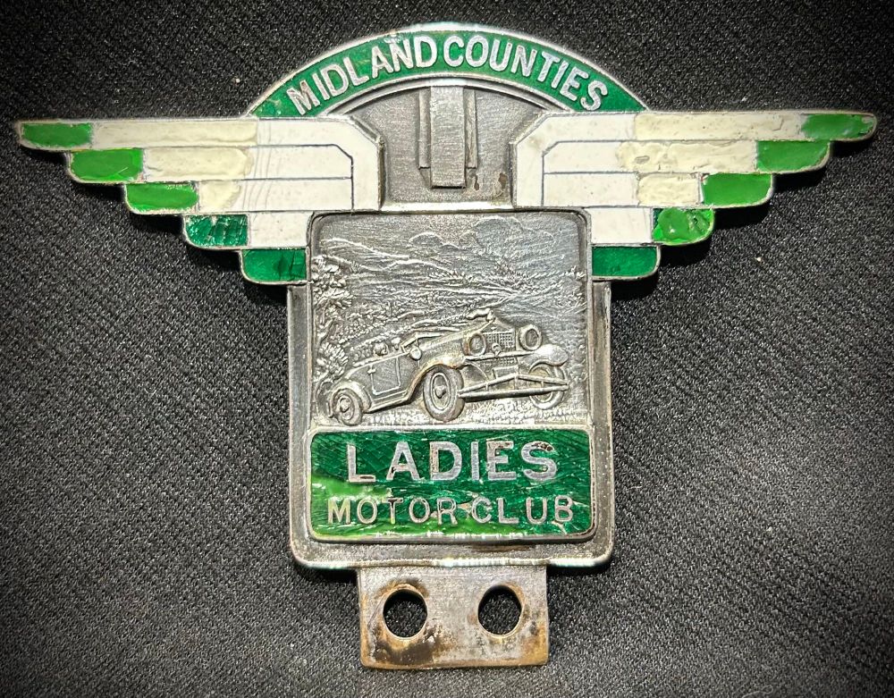 Nickel and green enamelled badge with “Midland Counties” wording in an arch of green enamel above deco green tipped white wings below which is an embossed panel depicting a late 1920’s/early 1930’s car climbing a hill at speed in a landscape of hills and clouds. Below is the wording “Ladies Motor Club” on a green enamel background.  The badge has a tang with two holes for badge bar mounting. The badge is 9cm wide and 7.5cm tall and has had some paint repairs to damaged enamel over its c.92 year life.
