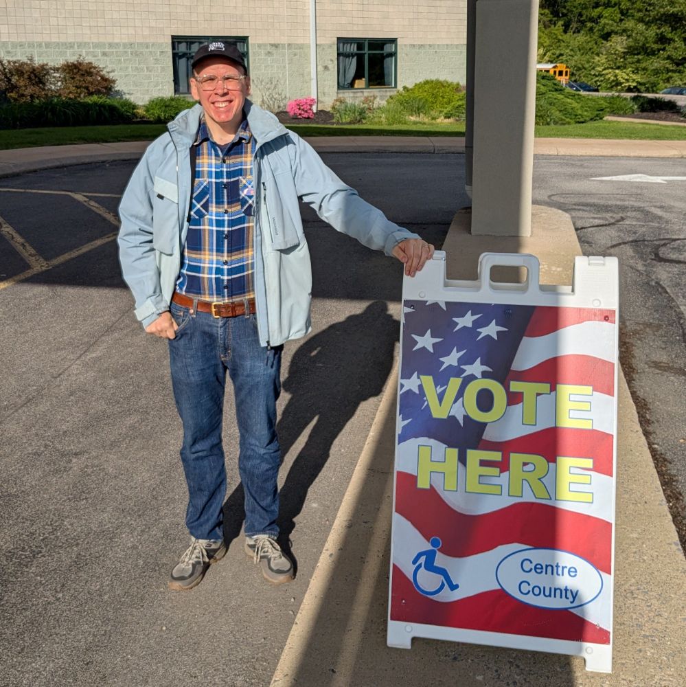 Matt Herndon wearing a black Vote Today PA hat and a blue coat, flannel shirt and jeans standing next to a Vote Here Centre County sign.