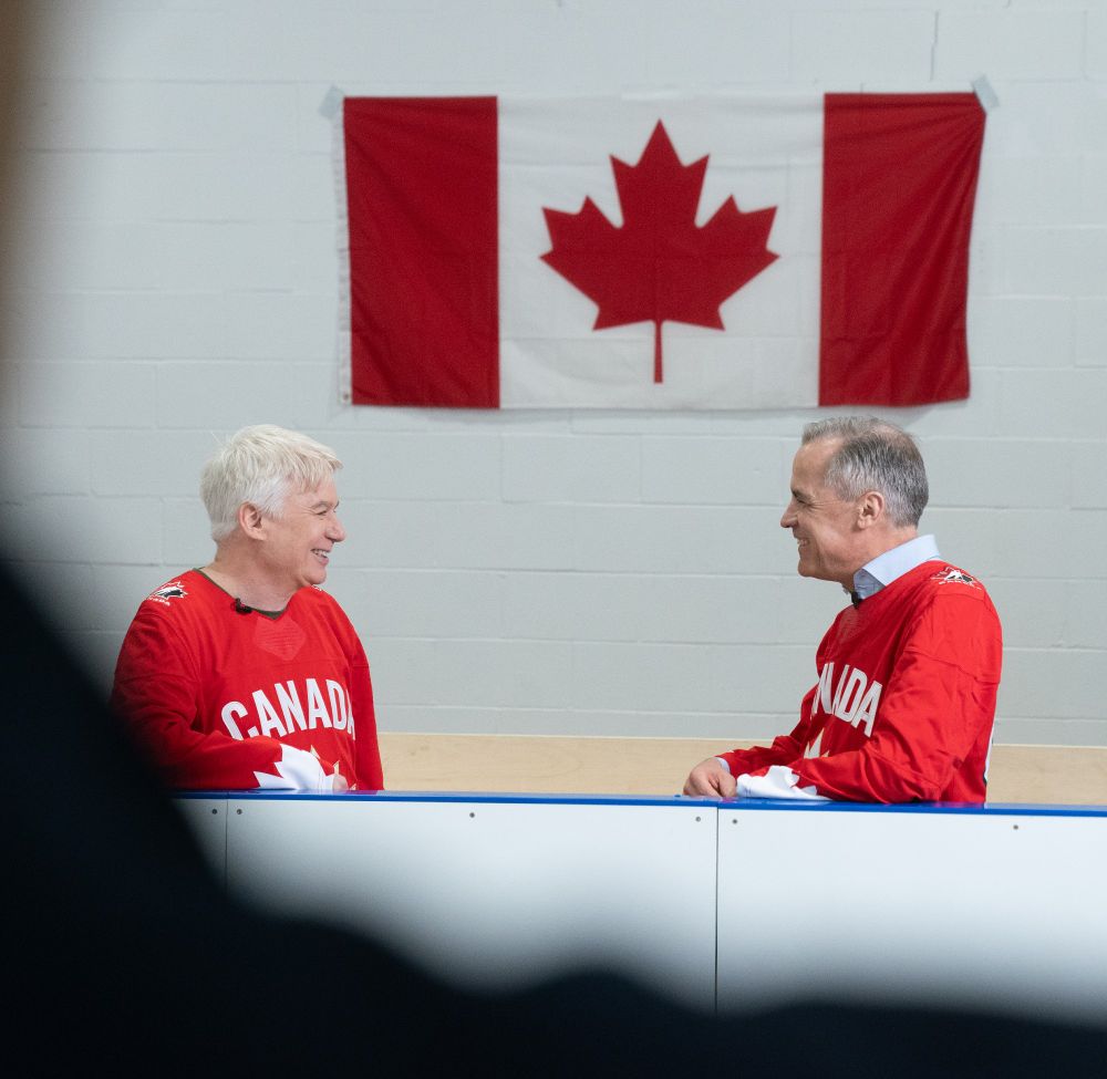 Mike Myers and Prime Minister Mike Carney are photographed standing together, leaning on the boards of an ice rink. They are talking to each other and smiling.