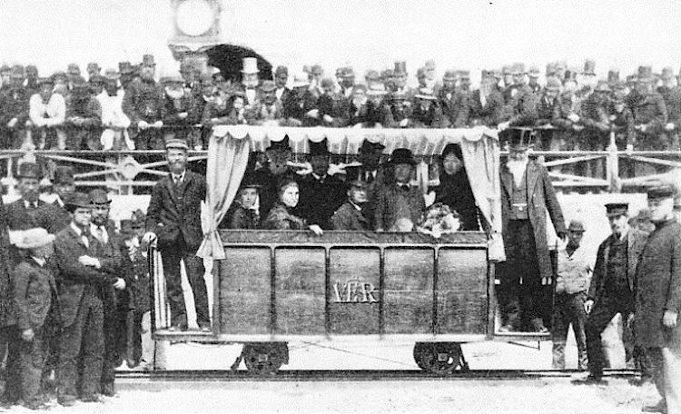 Monochrome photo of an open carriage on the beach, curtained, full of passengers (must be VIPs), with many people watching, all the men in hats!

