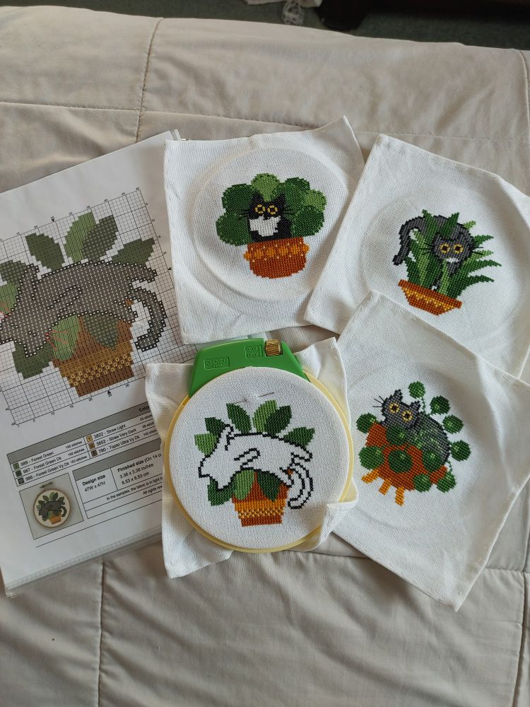 Four cross stitch projects of black and gray cats peeking out of green houseplants in brown pots