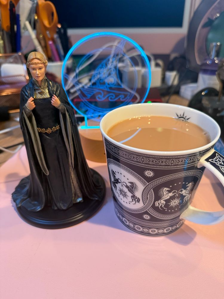 A small sculpture of Éowyn next to a cup of tea in a Rohan mug on a messy desk. 