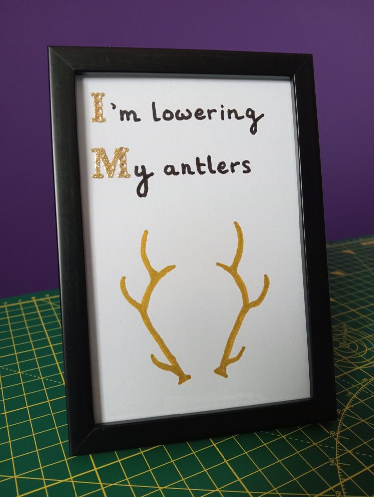 A black frame, with white card inside, sitting on a green cutting board in front of a purple wall. "I'm lowering my antlers" is written at the top, with I and M in sparkly gold and the rest in black. Underneath is a drawing of two antlers, in gold.