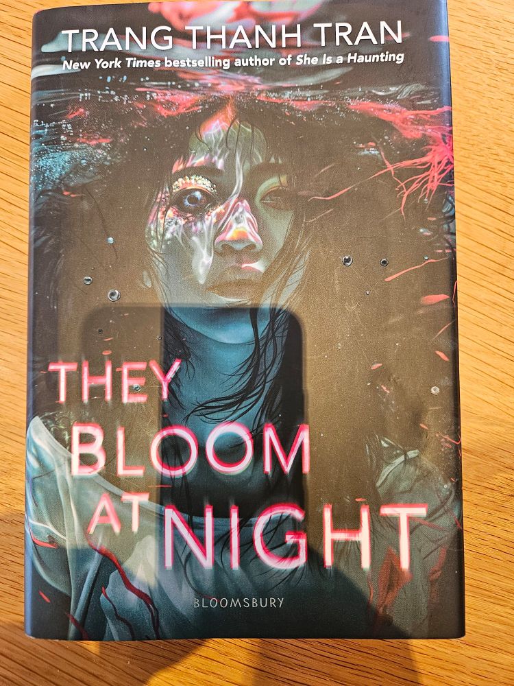 A picture of the cover of They Bloom at Night by Trang Thanh Tran. It shows a human teen transforming into an ocean being while floating just beneath the surface of water.