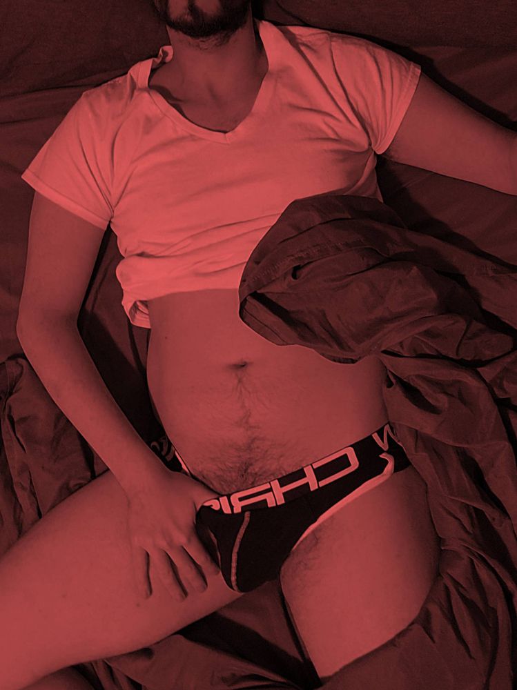 Deedee lounging on their bed in a jockstrap. The photo is tinted red.