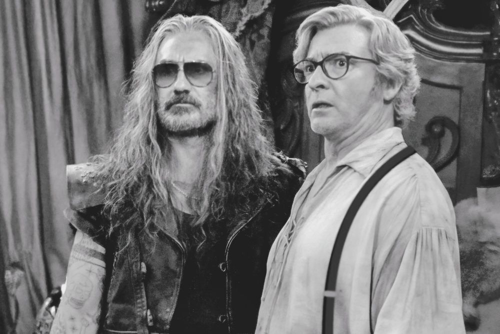 Black and white image of Ed and Stede standing next to each other, staring off to the left of the image in confusion. Ed is wearing his classic leather jacket with a pair of dark sunglasses superimposed over his eyes. Stede is wearing his billowing white shirt, a pair of drawn on suspenders, and a pair of dark rimmed glasses superimposed over his eyes.