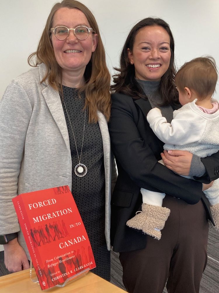 Professor Christina Clark-Kazak poses on the left, with Dr. Yuriko Cowper-Smith on the right (holding her baby), at the launch of the edited volume, "Forced Migration in/to Canada". The newly published book is on the counter in the foreground.
