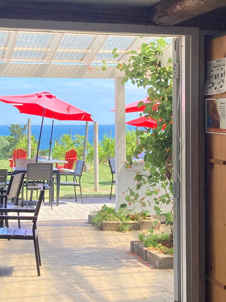 A view through a doorway of patio tables in the sunshine with red parasols providing a little shade. The blue banks of the Bay of Quinte in the background.