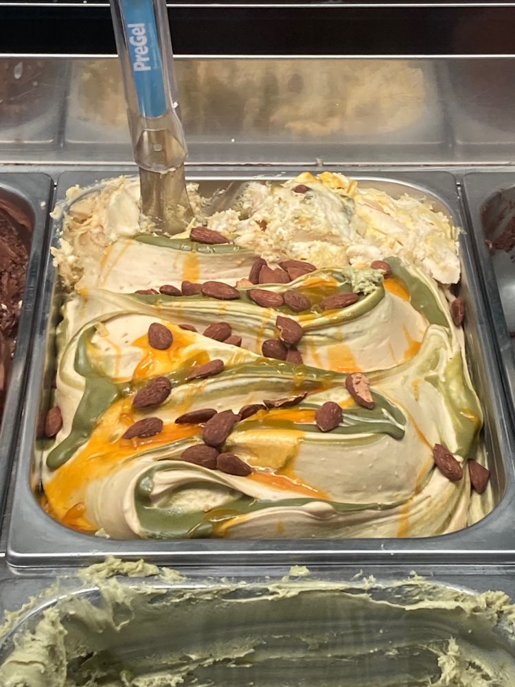 A light brown gelato in a metal tub with green and orange swirls atop and pieces of whole almonds.