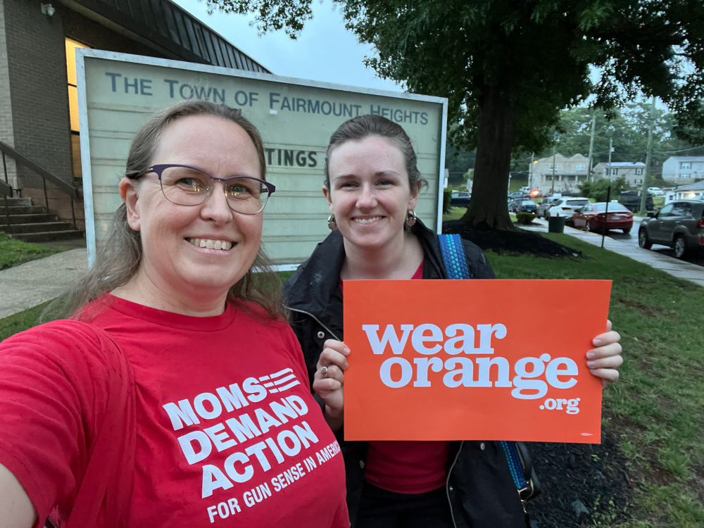 Two women in front of the Town of Fairmount Heights sign, one wearing a red shirt that says “Moms Demand Action for gun sense in America” and the other with an orange sign that says WearOrange.org.