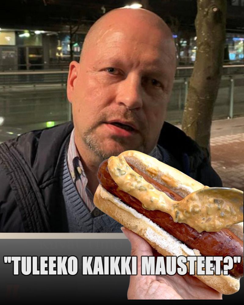 Timo Vornanen väsyneen näköisenä katsoo nakkikioskin myyjää, josta näkyy kuvassa vain käsi, jossa hotdog, jota maustaa parhaillaan. Teksti: Tuleeko kaikki mausteet?