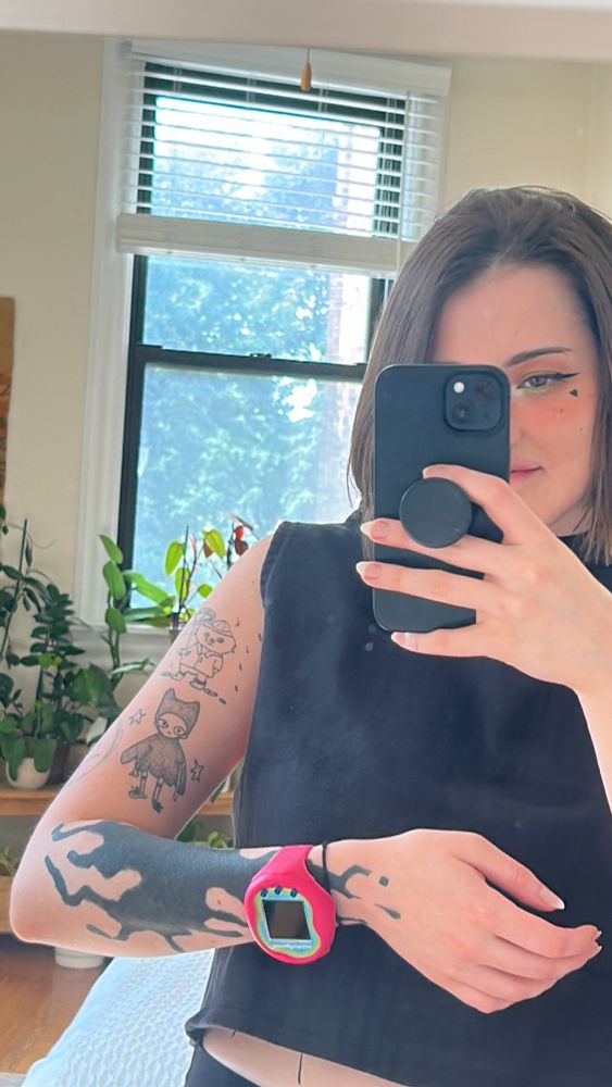 picture of me in a mirror smiling behind my phone, my arm is held across my chest and shows tattoos with a tamagotchi uni on my wrist. there is a large window in the background