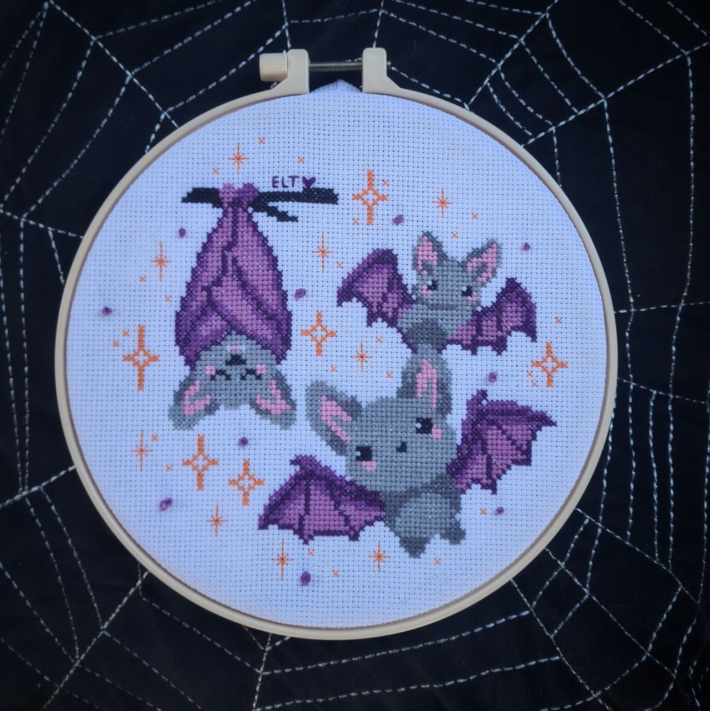 Cross stitch hoop featuring three cute cartoony bats, one sleeping hanging from a branch and the other two flying. Lots of star sparkles surround them. 