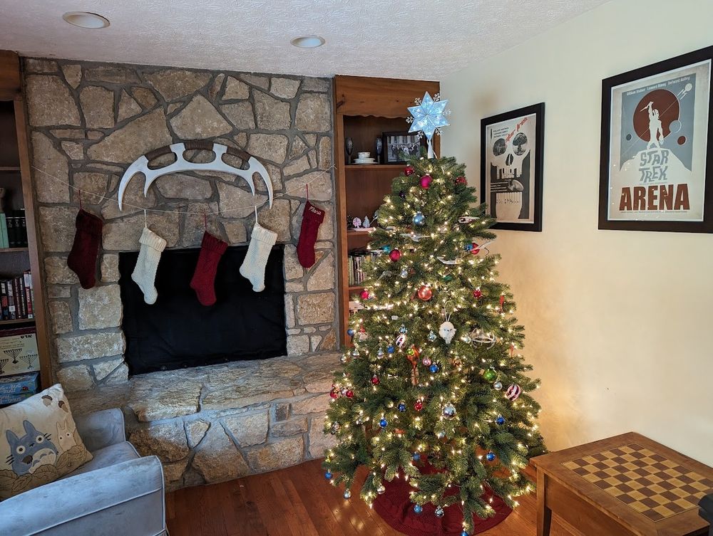 A lit Christmas tree with a 6 pointed star atop it standing beside a stone hearth across which are strung five knit stockings and above which hangs a large curved Klingon sword of honor. 