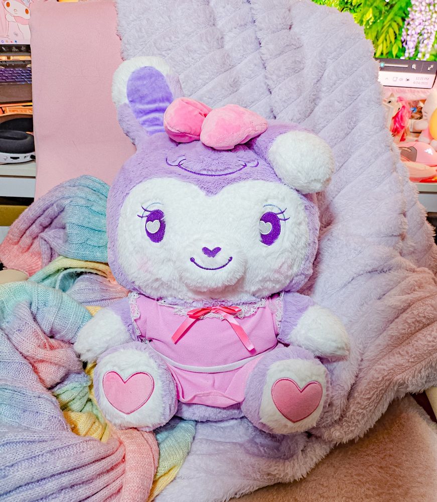 A stylized bipedal mostly purple and white bunny character plushie wearing a pink dress and sitting on a pink computer chair. A purple fuzzy blanket and rainbow fabric are behind her to add to the photo.
