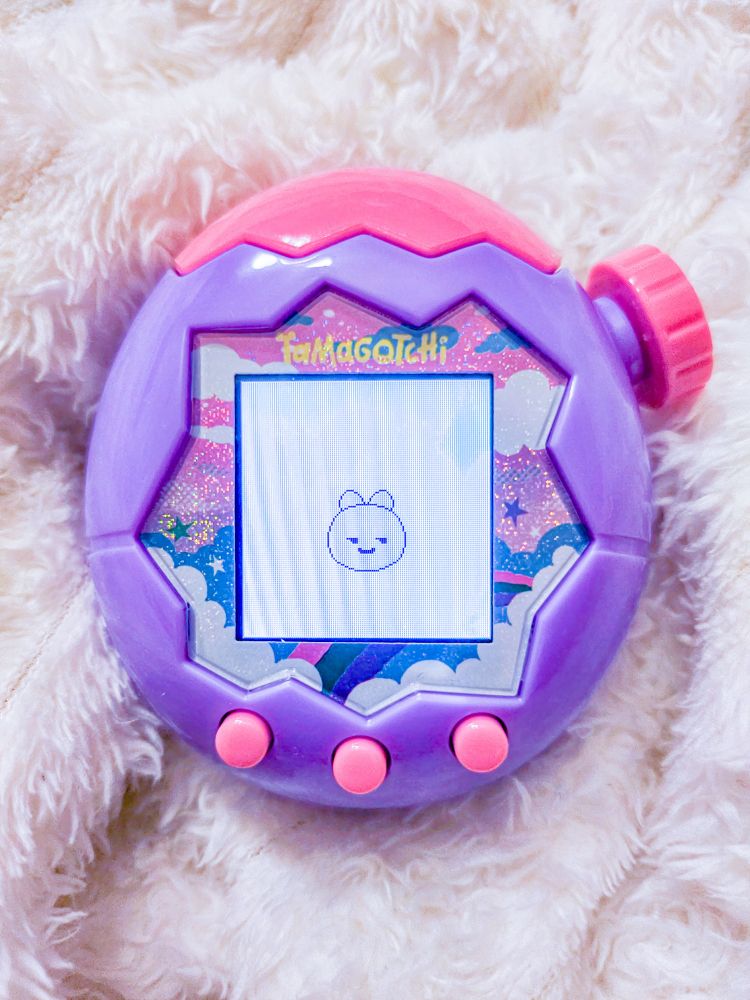 A purple and pink "Tamagotchi Paradise" Sky version with a blob-looking Tamagotchi character on the screen. The device is on a fluffy white blanket.
