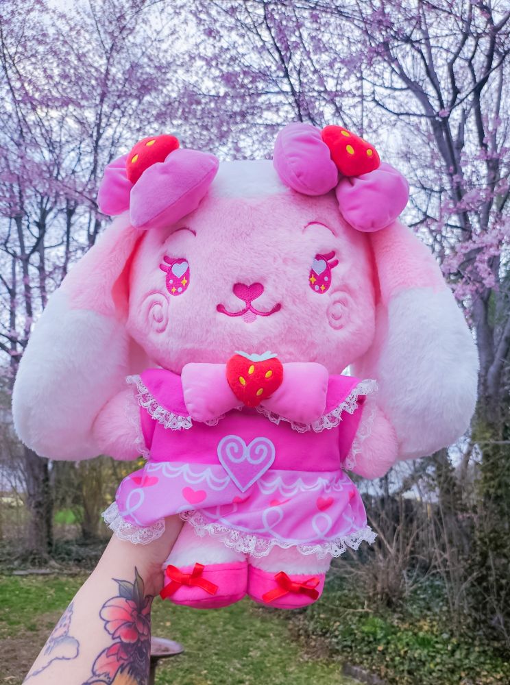 A stylized bipedal mostly light pink and white lop bunny character plushie wearing an ornate pink dress with strawberry embellishments and bows. She's held out against pink trees and green grass/shrubbery by a tattooed arm.