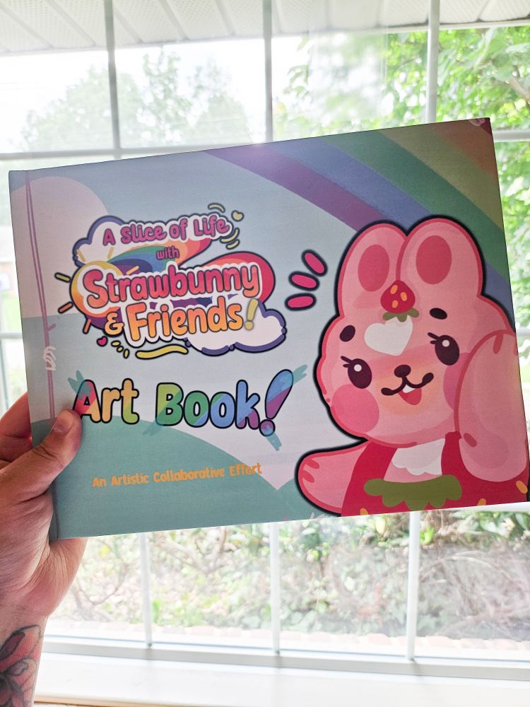 A book with a pink bunny in red overalls on the cover. The logo and text read, "'A Slice of Life with Strawbunny and Friends' Art Book!"
