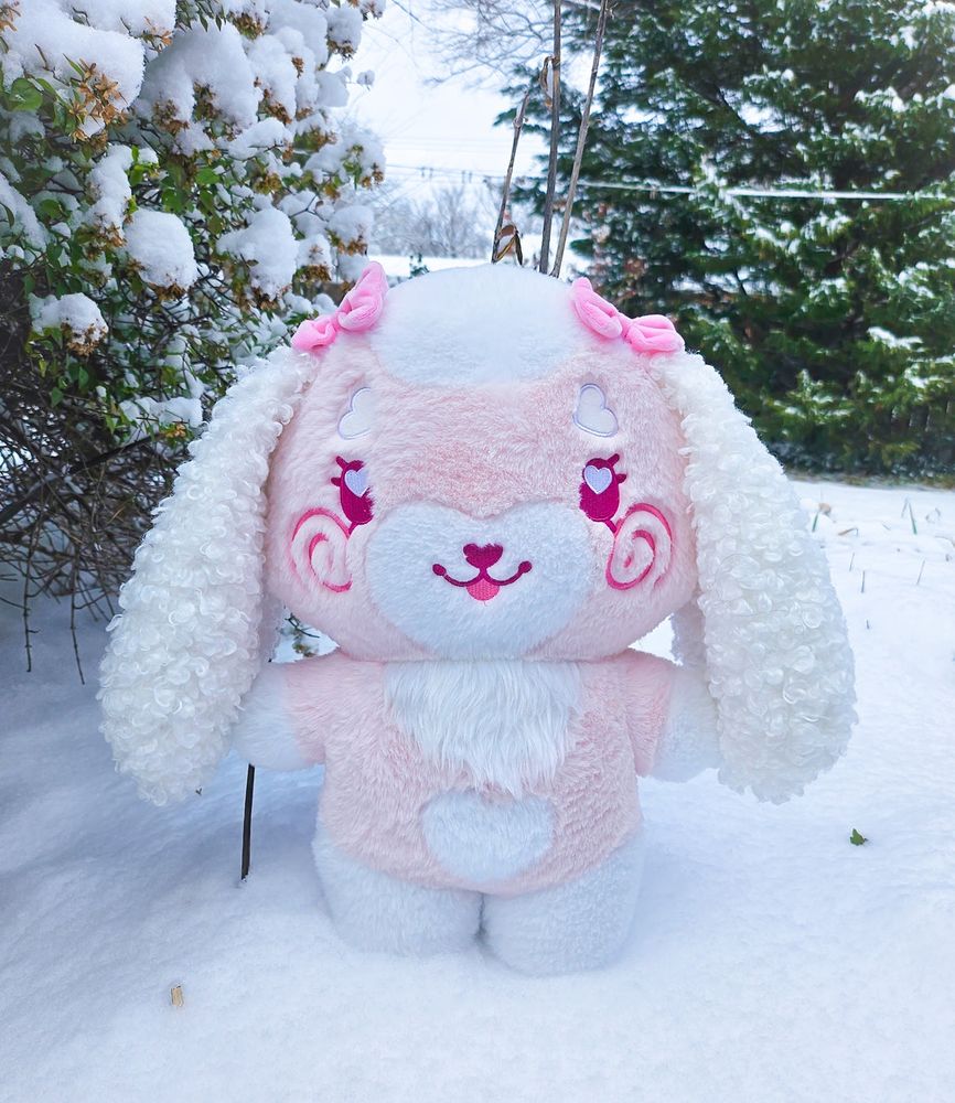 A stylized bipedal mostly light pink and white lop bunny character plushie standing in the snow