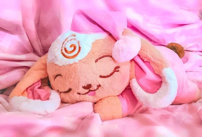 a stylized bipedal mostly beige lop bunny plush with white accents as bangs and the tips of her long ears. she's styled to appear sleeping in a lying down position with pink pajamas and a pink night cap on her head