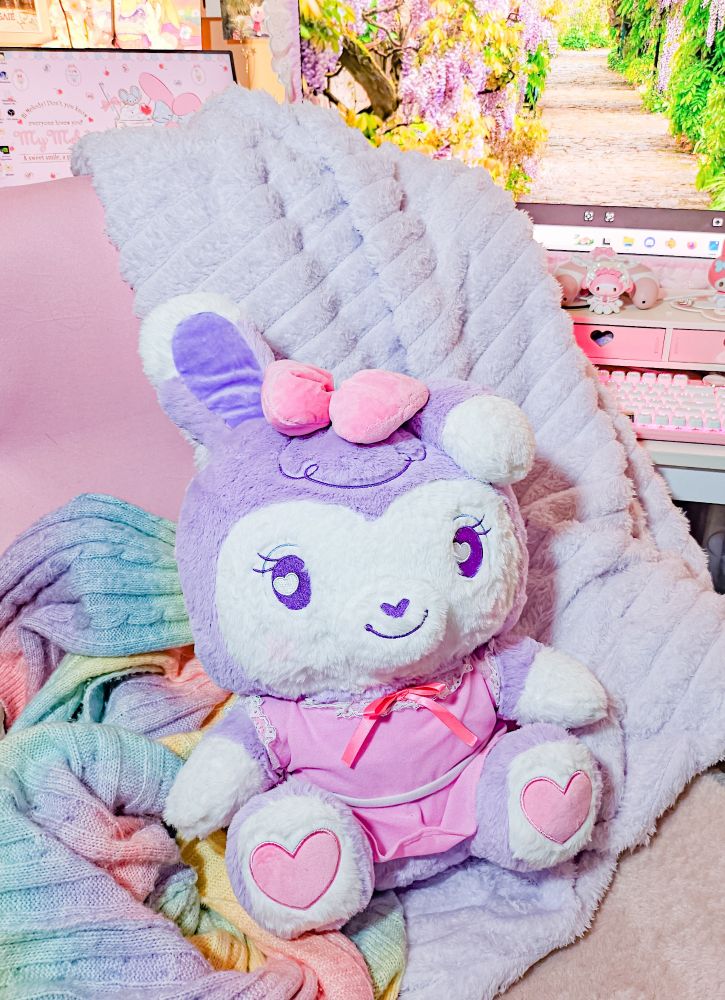 A stylized bipedal mostly purple and white bunny character plushie wearing a pink dress and sitting on a pink computer chair. A purple fuzzy blanket and rainbow fabric are behind her to add to the photo.

In the background is a desktop monitor with a Wisteria image displayed on it and a pink keyboard/gaming setup.