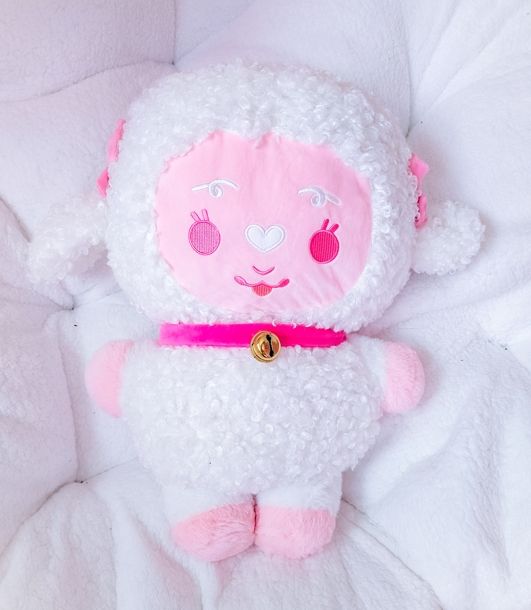 A stylized bipedal mostly white and light pink lamb plush with a pink bell collar sitting on a white cushion