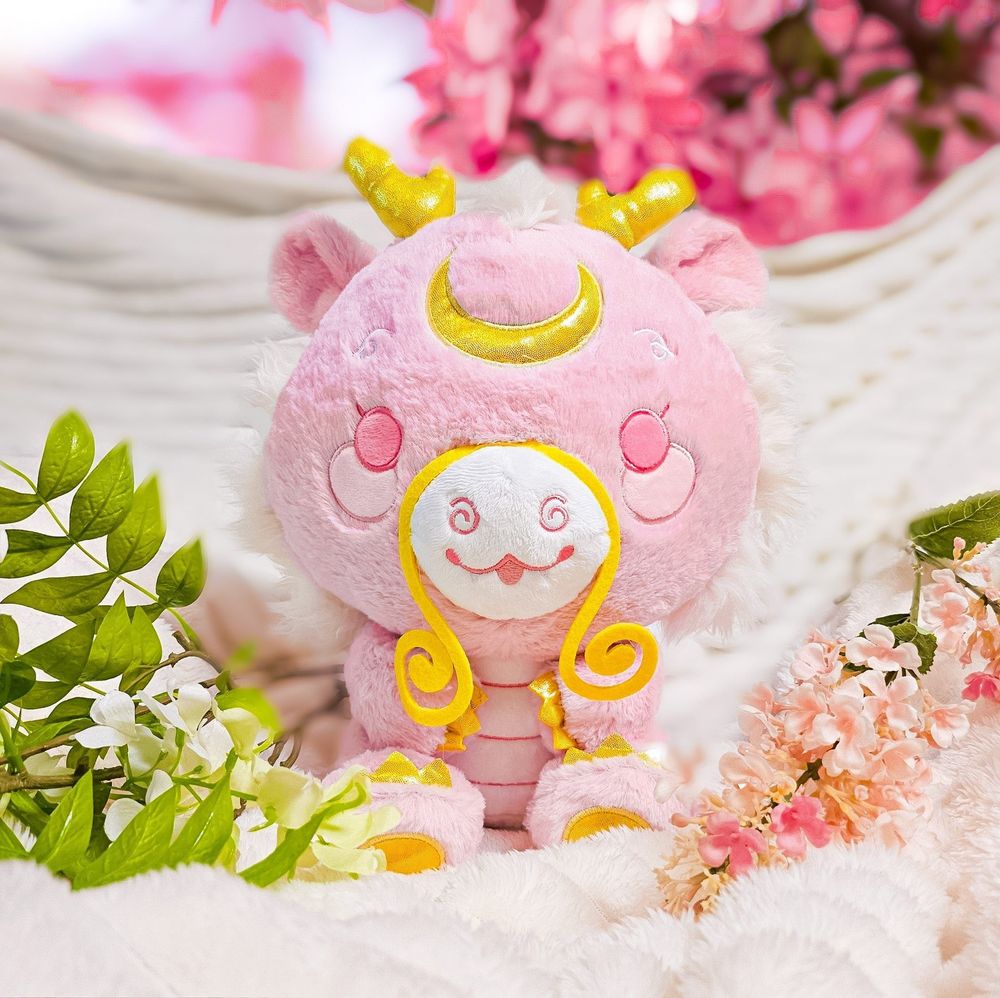 A mostly pink Chinese dragon bipedal character plushie with gold horns ans "whiskers" with a sparkly crescent Moon on his forehead. He's sitting amongst flower and leaves with a white blanket behind him.