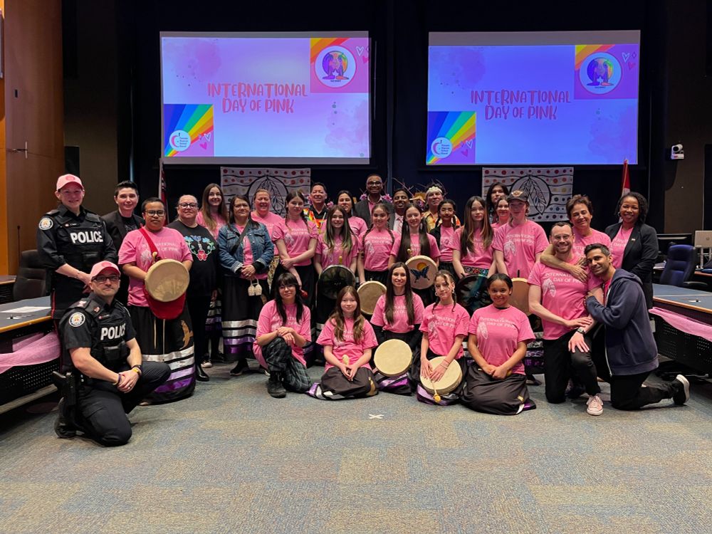 A group of people, including children and adults, are gathered in a conference room wearing pink shirts. Behind them, there are two large screens displaying the text 'INTERNATIONAL DAY OF PINK' with colourful backgrounds and logos.