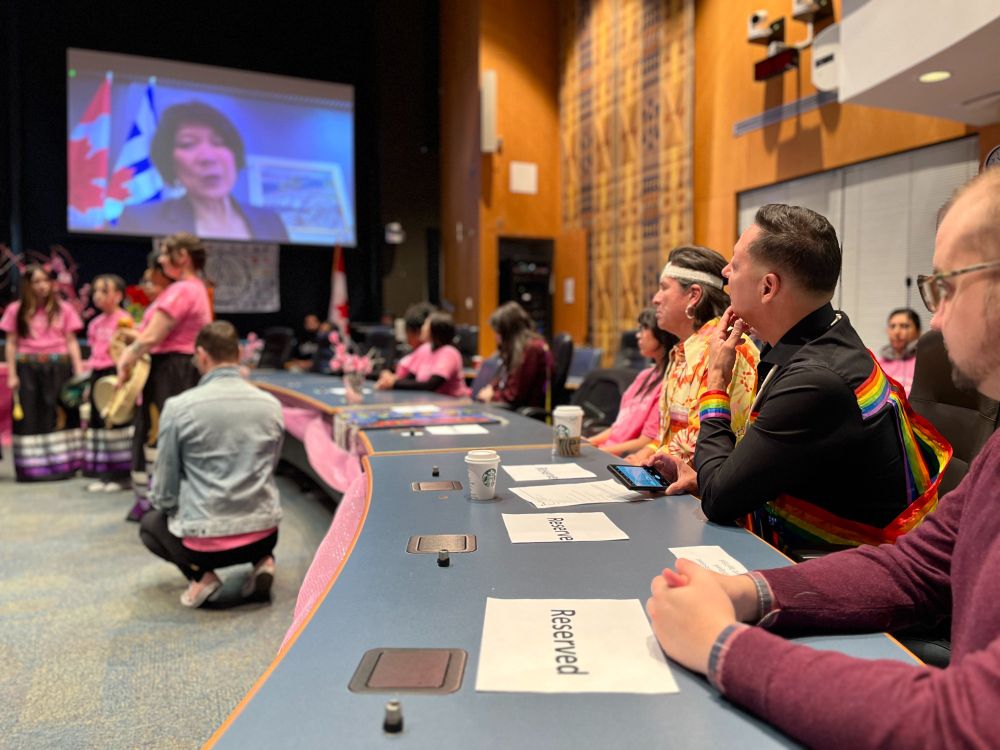 A group of people is seated in a conference room, watching a presentation on a large screen at the front.