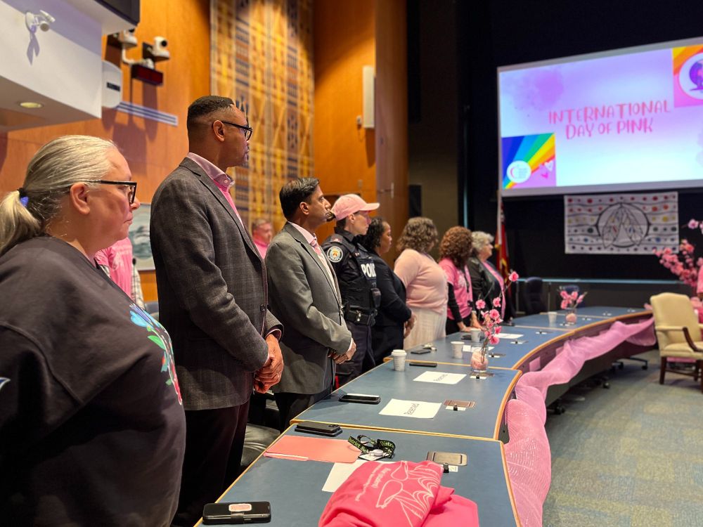 A group of people are standing in a conference room, facing a stage with a large screen displaying 'International Day of Pink.' 