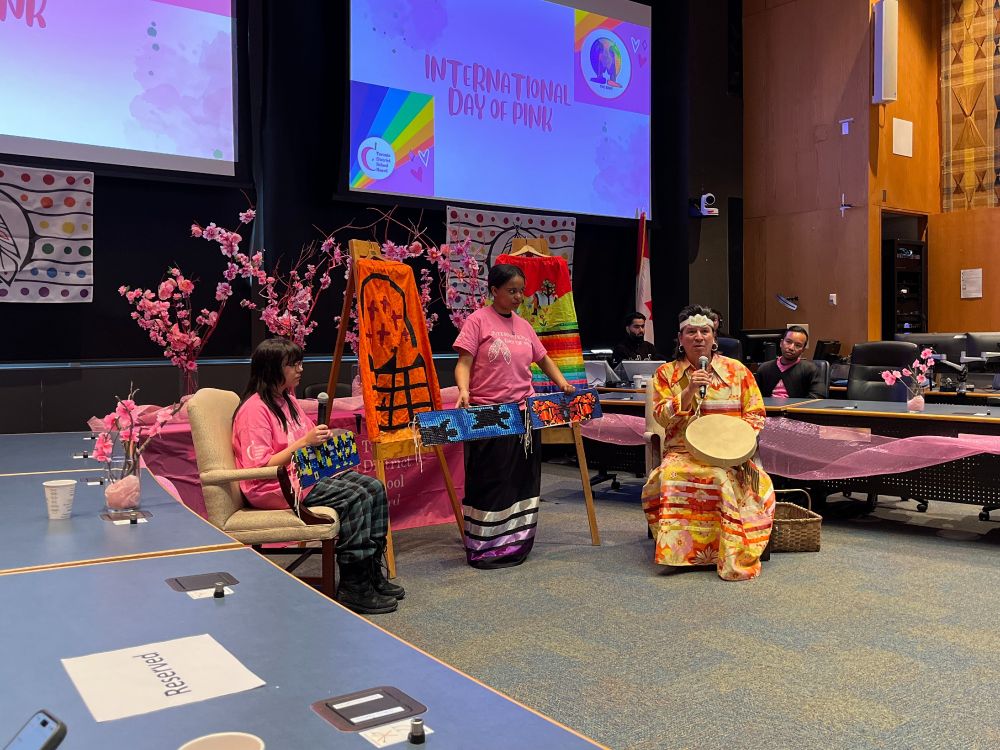 A group of people in stand in a conference room with two screens displaying 'International Day of Pink.' The room is decorated with pink-themed decorations.