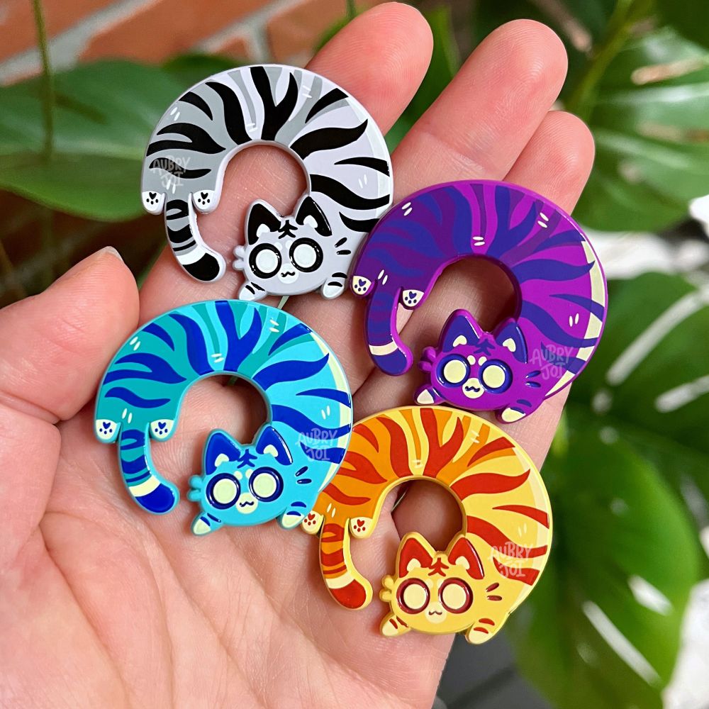 Four enamel pins of long cats in circular curled poses, designed to look like tigers. They’re each a different color theme: grayscale, magenta/purple, teal/blue, and orange.