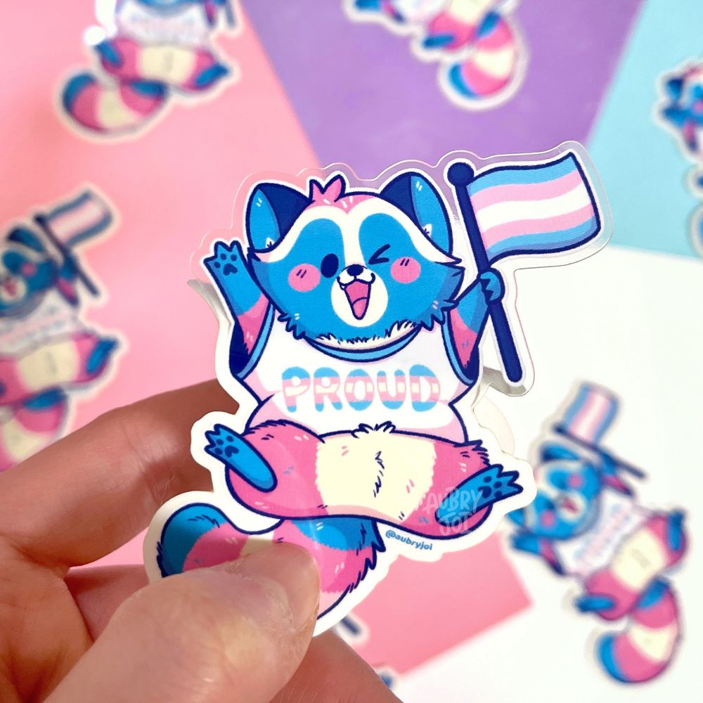 A sticker of Egg the Trans Raccoon standing with a silly expression, with text that says โtransition goals, just a little guyโ in glitter.