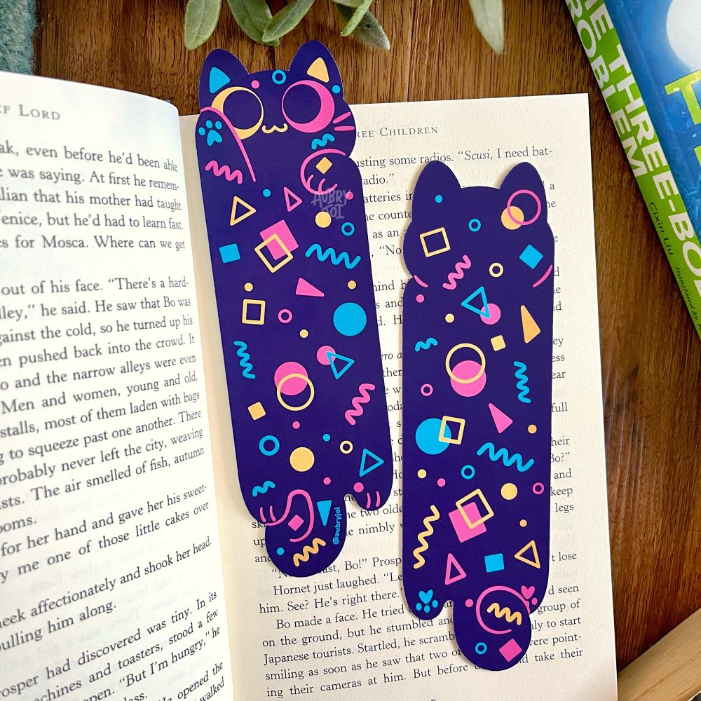 An open book showing off the front & back design of a diecut bookmark of a retro arcade carpet long cat.