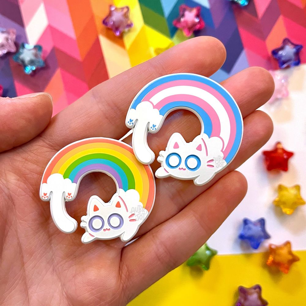 A hand holding two enamel pins of long cats curled into a circular pose. Each cat has a cloud themed head & tail with a rainbow shown along their bodies. One is standard rainbow colors, and the other is trans flag colors.