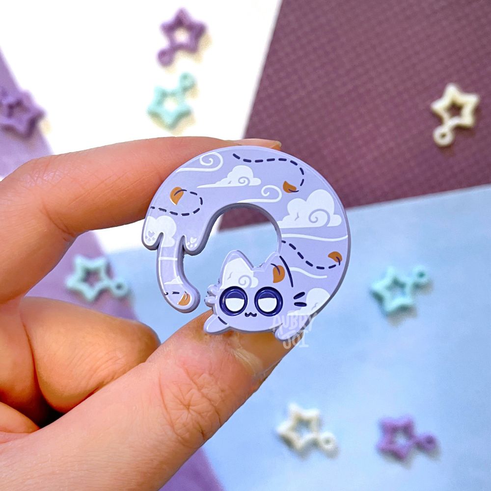 A hand holding an enamel pin of a long cat curled into a circular shape. The cat is a pale purple/periwinkle with a motif of clouds, windy movement lines, and small leaves across it. Its eyes are half-lidded and lazy looking.