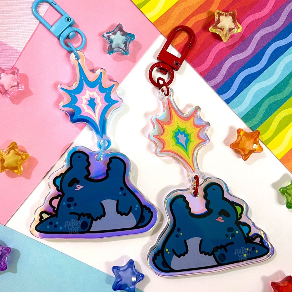 A photo of two keychains of kawaii-style, chubby Godzillas with their heads tilted back and flames bursting from their open mouths. The flames are separate, connected pieces of acrylic, and one is  colored blue, pink & white like the trans flag; the other is colored rainbow. There are star charms and colorful paper in the background.