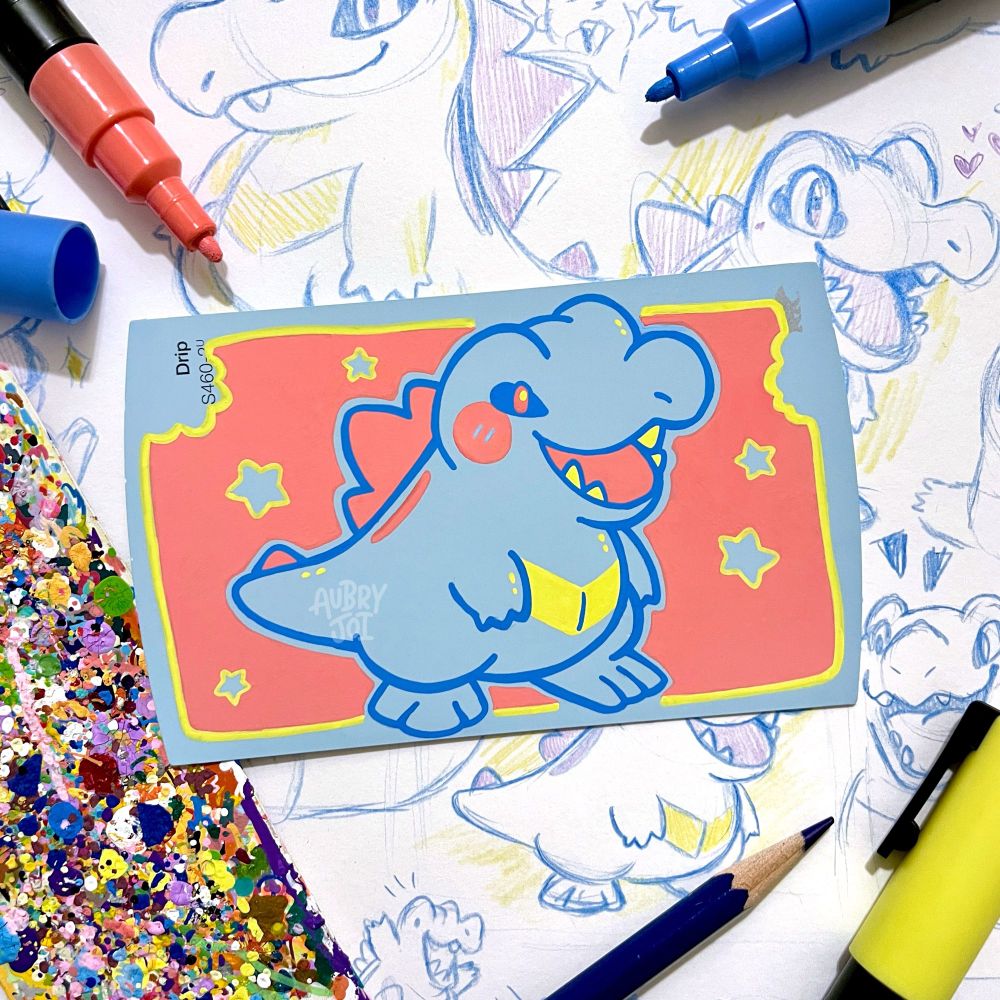 A cute, stylized paint pen illustration of Totodile on a blue paint swatch card. The color of the card is used as Totodile’s base, so he’s mainly illustrated with outlines. The painting sits on top of a sketchbook page filled with Totodile sketches.