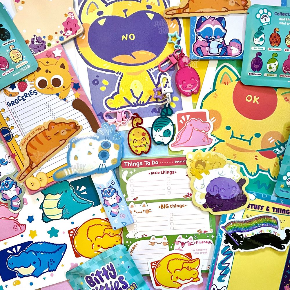 A spread of merchandise by Aubry Joi, to celebrate their July shop update. Everything is very cute and colorful, and mostly features cats. There are blind bag acrylic keychains, notepads & sticky notes, many stickers, and art prints.
