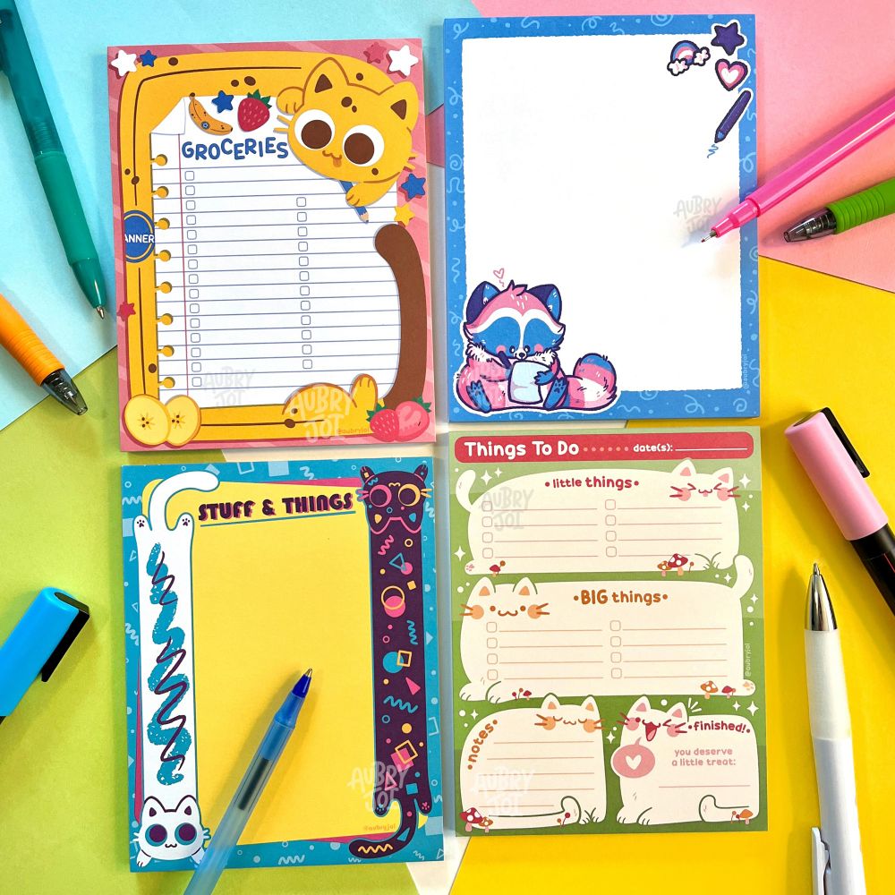 Four unique notepad designs by Aubry Joi. Each is cute & colorful with stylized characters on it. They are of: a banana long cat making a grocery list; Egg the Trans Raccoon writing a note; retro pattern-themed long cats; chubby cats making up a to-do list. Pens are scattered around the pads.
