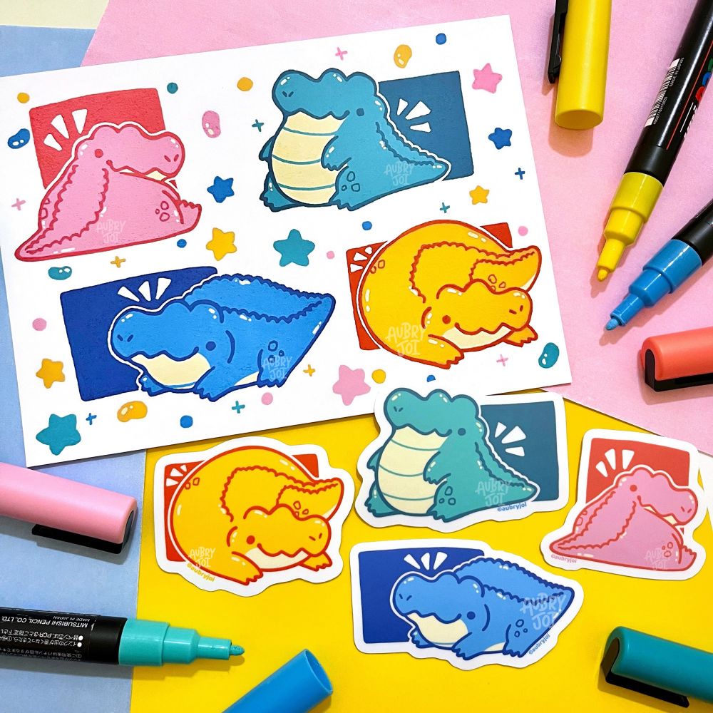 A spread of items featuring chubby, cute pastel colored crocodiles. The crocodiles are colored: pink, teal, blue, yellow. There’s an art print showing all 4 crocs, as well as a set of stickers, one of each croc.