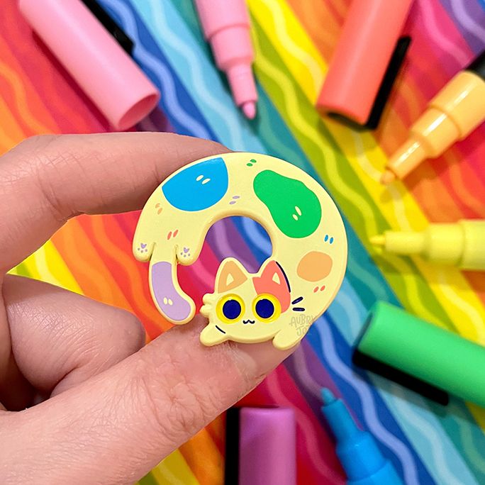 A photo of a hand holding an enamel pin of a long cat curled into a circular pose. The cat is a soft cream color with pastel rainbow patches and big, yellow eyes. The photo is taken over a colorful paper with Posca pens around it.