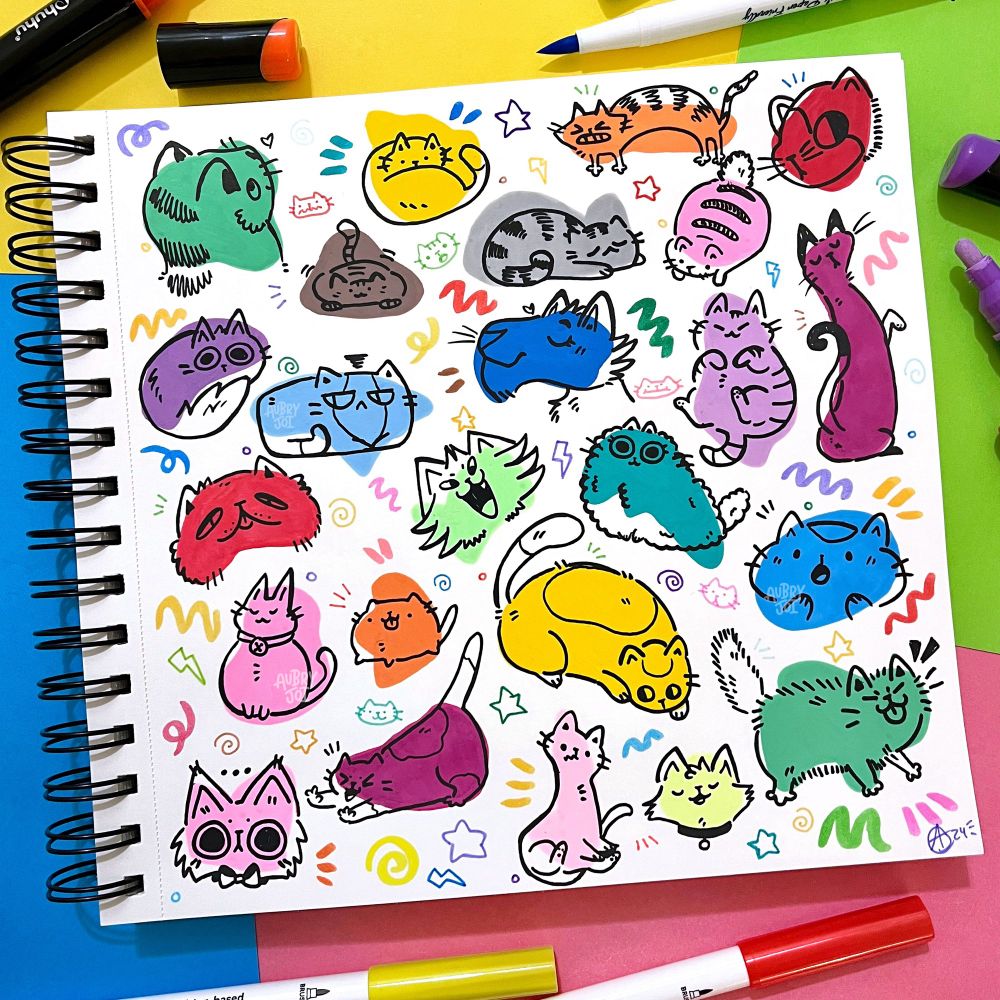 A sketchbook open to show a page full of colorful blobs with black lineart over them to turn them into cats. Each is unique and there are tiny marker doodles between them. The blobs were painted with paint pen, and a few lay around the book on colorful paper.