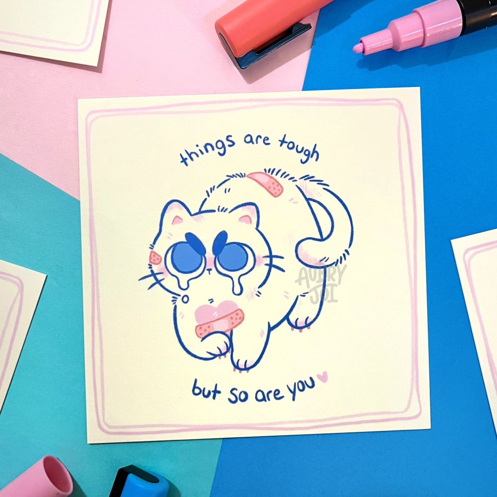 An art print of a slightly beat-up looking white cat with huge, watery angry eyes. They have a couple of bandages on them, one placed over a pink heart marking on their chest, and are in a defensive pose. Around them it says “things are tough, but so are you.” 